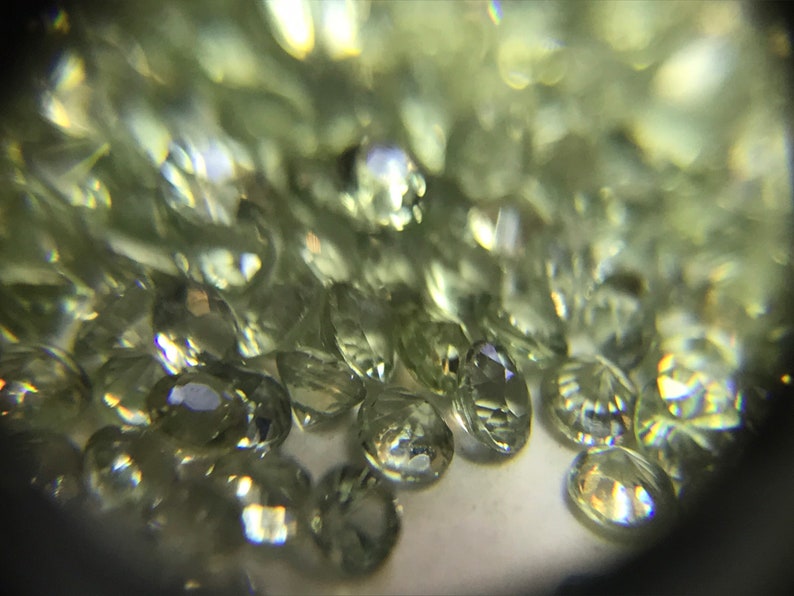May include: A close-up view of a pile of small, faceted, light green gemstones. The gemstones are oval-shaped and have a shiny, reflective surface.