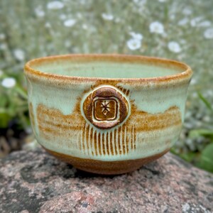 May include: A small, light green ceramic bowl with a brown glaze and a decorative design. The bowl has a brown rim and a brown, star-shaped design in the center.