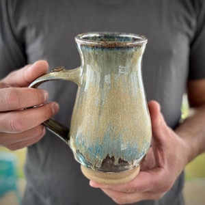 Può includere: Una tazza in ceramica fatta a mano con una glassa strutturata unica nei toni del blu, beige e marrone. La tazza ha un manico curvo e un bordo ampio e svasato. La tazza è tenuta da una persona.