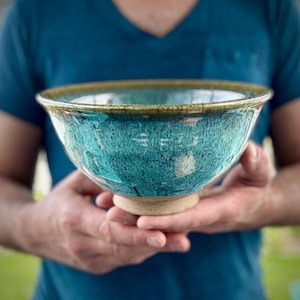 May include: A large, teal-colored ceramic bowl with a brown rim and a speckled glaze. The bowl is sitting on a wooden base.