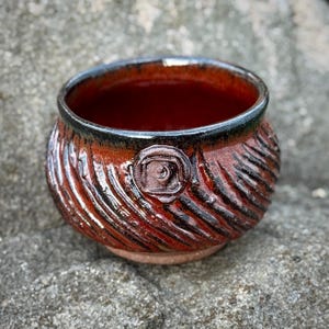 May include: A handmade ceramic bowl with a textured, ridged exterior in shades of red and black. The rim is dark, and a circular emblem is centered on the side. The bowl is likely used for serving or display.