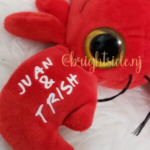 K&ouml;nnte beinhalten: Roter Pl&uuml;sch-Hummer mit Glitz Augen und den Namen "JUAN & TRISH", die auf eine Kralle gestickt sind.