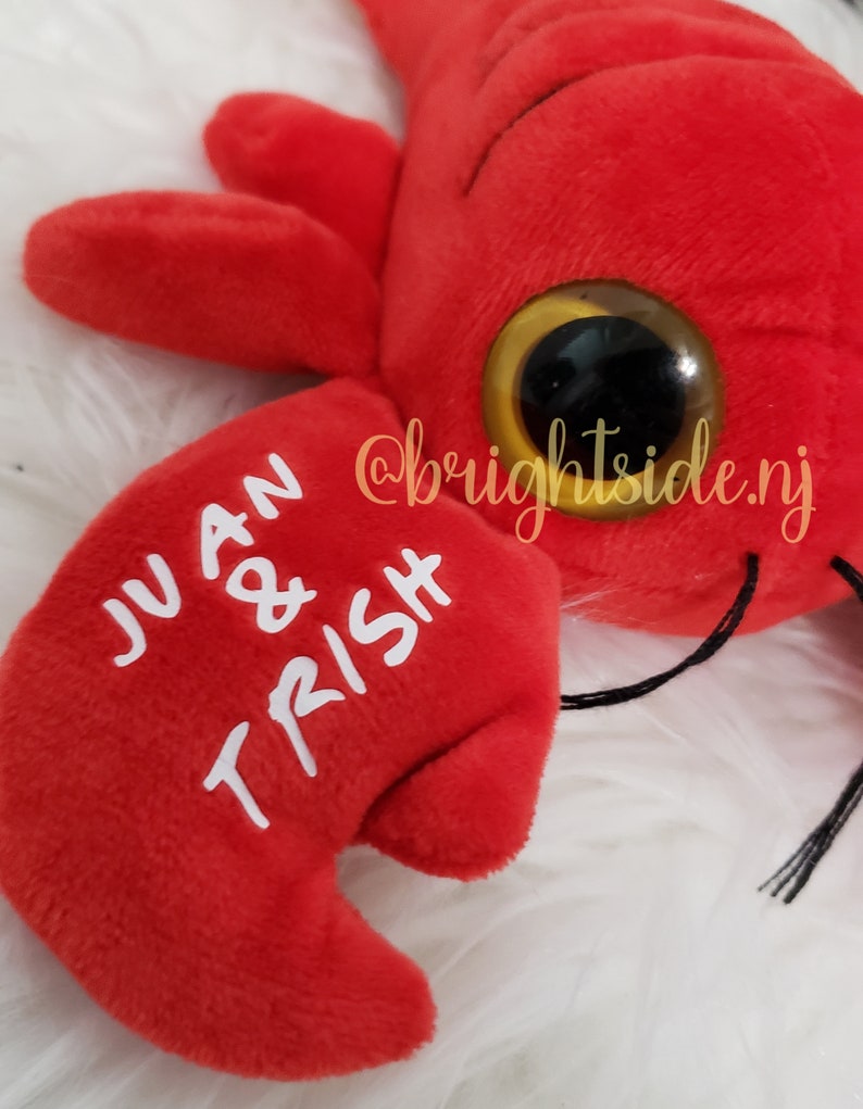 K&ouml;nnte beinhalten: Roter Pl&uuml;sch-Hummer mit Glitz Augen und den Namen "JUAN & TRISH", die auf eine Kralle gestickt sind.