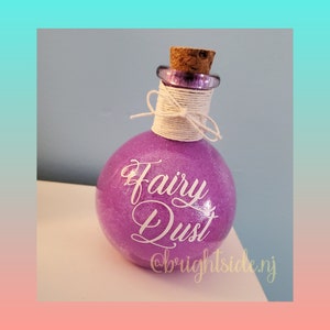 May include: A small purple glass bottle with a cork stopper and white twine tied around the neck. The bottle is filled with glitter and has the words "Fairy Dust" written on it in white lettering. The bottle also has the text "@brightside.nj" written on it.