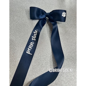 Puede incluir: Un lazo de satén azul marino con una huella de pata blanca y el texto "Penn State" impreso en un lado. La cinta está atada en un lazo y tiene dos colas largas. El texto "@BRIGHTSIDE.NJ" está impreso en la parte inferior de la imagen.