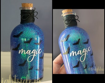 May include: A blue glittered glass bottle with black bat silhouettes and the word "magic" in white script. The bottle has a cork stopper and is wrapped with black twine, creating a Halloween or witchy theme.