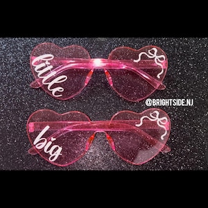 May include: Two pairs of pink heart-shaped sunglasses with glitter. One pair says "little" and the other says "big".