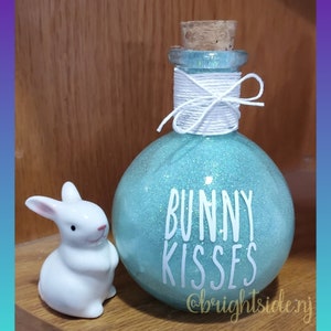 May include: A round, turquoise glitter-covered glass bottle with a cork stopper and white twine tied around the neck. The bottle has the words "BUNNY KISSES" in white. A small white ceramic bunny figurine is next to the bottle.