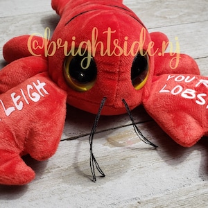K&ouml;nnte beinhalten: Roter Pl&uuml;sch-Hummer mit Glitz Augen und dem Text "You're my lobster" auf einer Kralle und "Ashleigh" auf der anderen Kralle.
