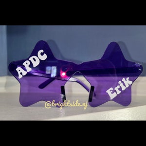 May include: Purple star-shaped sunglasses with the text "APDC" on one lens and "Erik" on the other lens.