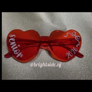 May include: Red heart-shaped sunglasses with glittery lenses. The text "Senior 2026" is written in white script across the lenses. The sunglasses have a clear bridge and arms. The background is a sparkly silver.