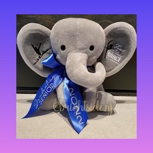 May include: A gray plush elephant with blue ribbon bows. The ribbon on the left side says "Graduating Senior 2022" and the ribbon on the right side says "First Class Dance Academy 2022".