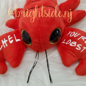 K&ouml;nnte beinhalten: Roter Pl&uuml;sch-Hummer mit gro&szlig;en Kulleraugen und schwarzen Antennen. Der Hummer hat die Worte "Rachel" und "You're My Lobster" in wei&szlig; darauf geschrieben.