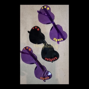 May include: Three pairs of heart-shaped sunglasses with different colored frames and lenses. One pair is purple with yellow stars, another is black with red stars, and the last pair is purple with yellow stars. The sunglasses have the words "tigers" and "rutgers" printed on them.