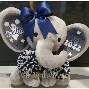 May include: Gray plush elephant with a blue bow, pom poms, and glitter accents. The elephant's ears are decorated with white stars and the text "Cecilia" and "2022".