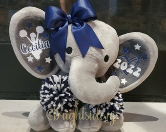 May include: Grey plush elephant with a blue bow, pom poms, and glitter accents. The elephant's ears are decorated with white stars and the text "Cecilia" and "2022".
