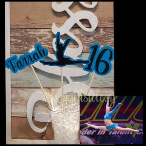 May include: A set of three blue and black glitter paper decorations with the words "Farrah" and "16" and a silhouette of a gymnast. The decorations are on wooden sticks and are in a clear glass vase with white shredded paper.