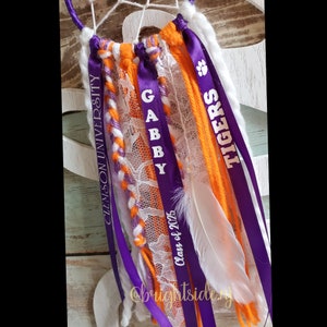 May include: Purple and orange ribbons with white lace and a white feather. The ribbons have the text "Clemson University", "Gabby", "Class of 2025", and "Tigers" printed on them.