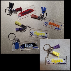 May include: Five clear acrylic keychains with colorful tassels. Each keychain has a different design with text and a heart. The designs include "tigers Farrah", "fcda little", "gators cheer", "tigers Farrah", and "rutgers".