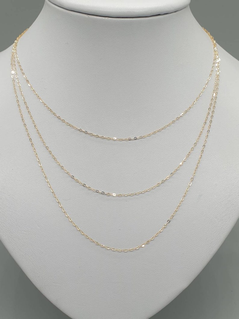 May include: A delicate gold chain necklace featuring three layered strands of fine, polished oval links. The necklace is displayed on a neutral-toned bust, showcasing the tiered arrangement of the thin, shimmering metallic chains that drape gracefully.