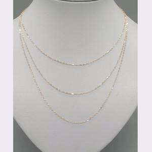 May include: A delicate gold chain necklace featuring three distinct layers of fine, shimmering links. The necklace is displayed on a neutral-toned bust, showcasing the tiered arrangement of the thin, polished gold metal strands that drape gracefully.