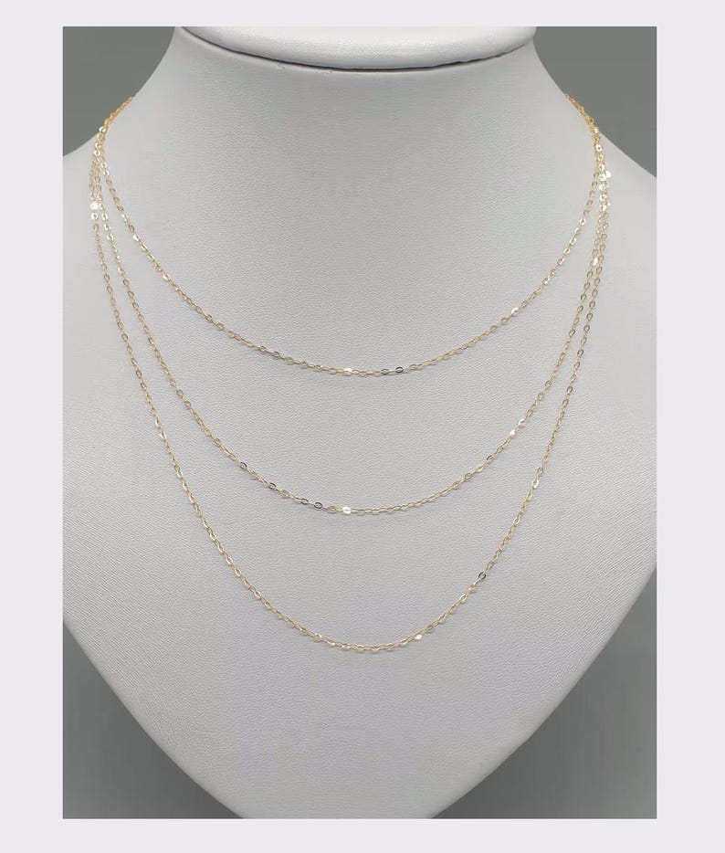 May include: A delicate gold chain necklace featuring three distinct layers of fine, shimmering links. The necklace is displayed on a neutral-toned bust, showcasing the tiered arrangement of the thin, polished gold metal strands that drape gracefully.