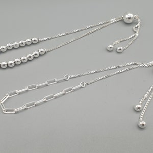 Puede incluir: Dos pulseras ajustables de plata. Una presenta una cadena de pequeñas cuentas redondas, y la otra tiene eslabones rectangulares. Ambas tienen un cierre de cuentas deslizante y detalles de bolas colgantes.