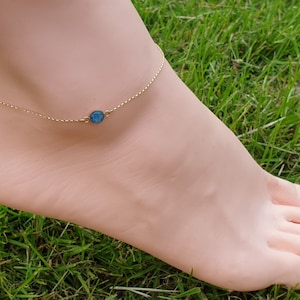 May include: A gold-colored anklet with a small, round, blue-green gemstone pendant. The delicate chain is draped around an ankle, with the foot resting on green grass. The gemstone is approximately 0.25 inches in diameter.