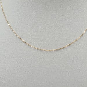 May include: A delicate gold chain necklace featuring small, uniform oval-shaped links. The chain is thin and polished, creating a continuous, refined metallic loop that hangs in a gentle curve, showcasing the fine craftsmanship of the metalwork.