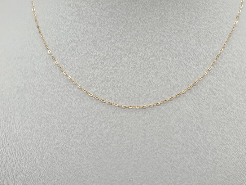 May include: A delicate gold chain necklace featuring small, uniform oval-shaped links. The chain is thin and polished, creating a continuous, refined metallic loop that hangs in a gentle curve, showcasing the fine craftsmanship of the metalwork.