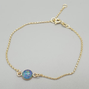 May include: A delicate gold bracelet featuring a round, iridescent opal stone. The opal displays shades of blue and green. The bracelet has a fine chain and an adjustable clasp. The jewelry is set against a neutral background.