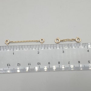 May include: Two gold-colored chain extenders with spring ring clasps. Each extender is approximately 1 inch long, with a chain section connecting the clasps. The extenders are displayed on a white surface next to a ruler.