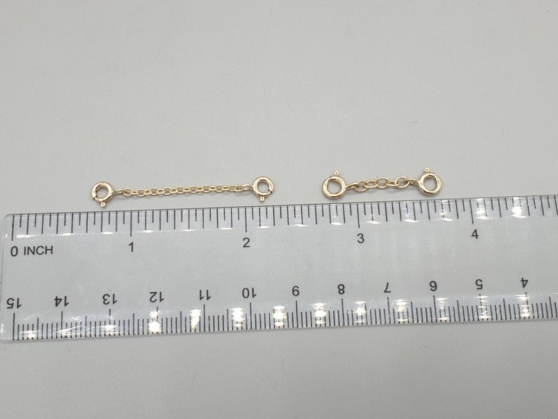 May include: Two gold-colored chain extenders with spring ring clasps. Each extender is approximately 1 inch long, with a chain section connecting the clasps. The extenders are displayed on a white surface next to a ruler.