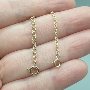 May include: Two gold-colored chain extenders with lobster claw clasps. One extender is longer than the other. The chains are made of small, linked oval rings. The clasps are round with a small lever to open and close.