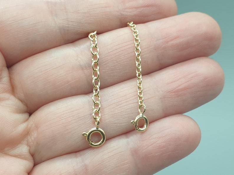 May include: Two gold-colored chain extenders with lobster claw clasps. One extender is longer than the other. The chains are made of small, linked oval rings. The clasps are round with a small lever to open and close.