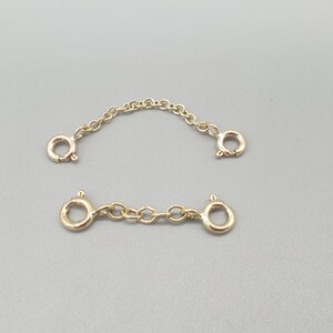 May include: Two gold-colored chain extenders with lobster claw clasps. One extender is a short chain, and the other is a longer, curved chain. The extenders are displayed on a gray background.