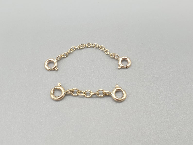 May include: Two gold-colored chain extenders with lobster claw clasps. One extender is a short chain, and the other is a longer, curved chain. The extenders are displayed on a gray background.