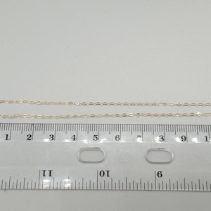May include: A delicate gold-toned metal chain necklace positioned above a clear plastic ruler. The ruler shows the chain segment measures approximately 11 centimetres in length. The chain features small, uniform oval links connected in a series.