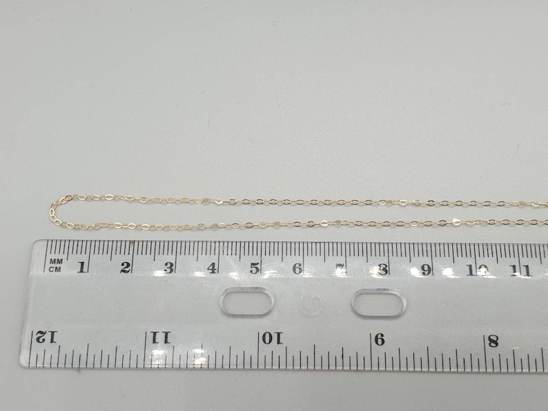 May include: A delicate gold-toned metal chain necklace positioned above a clear plastic ruler. The ruler shows the chain segment measures approximately 11 centimetres in length. The chain features small, uniform oval links connected in a series.