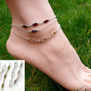 May include: Silver anklets featuring oval gemstones in shades of red, orange, and clear. The anklets are worn on a foot, set against a backdrop of green grass. Additional anklets with various gemstone colours are shown in the lower left.