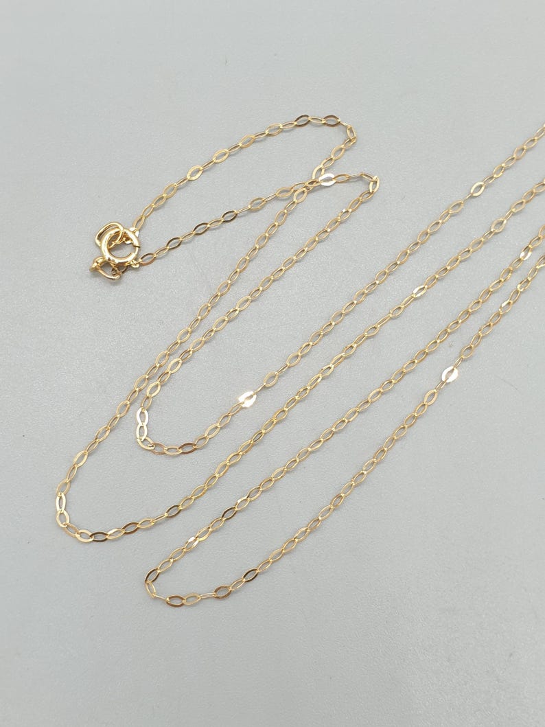 May include: A delicate gold chain necklace featuring elongated oval links. The necklace includes a small circular spring ring clasp at one end, allowing for secure fastening. The chain is composed of thin, polished gold metal links.
