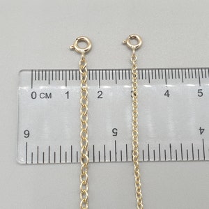 May include: Two gold-colored chain extenders with lobster claw clasps are displayed against a gray background next to a ruler. The chains are different lengths, with one measuring approximately 2 inches and the other approximately 1 inch.