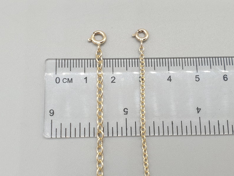 May include: Two gold-colored chain extenders with lobster claw clasps are displayed against a gray background next to a ruler. The chains are different lengths, with one measuring approximately 2 inches and the other approximately 1 inch.