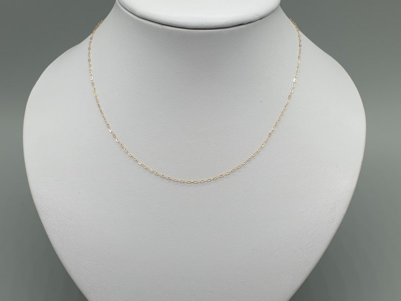 May include: A delicate gold chain necklace featuring small, uniform oval-shaped links. The fine metal chain is displayed in a simple, continuous loop, showcasing a polished, reflective surface that catches the light across its entire length.