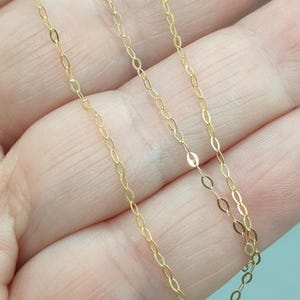 May include: A delicate gold chain necklace featuring a series of small, elongated oval links connected in a continuous pattern. The metal has a bright, reflective finish that catches the light, creating a subtle shimmer along the length of the chain.