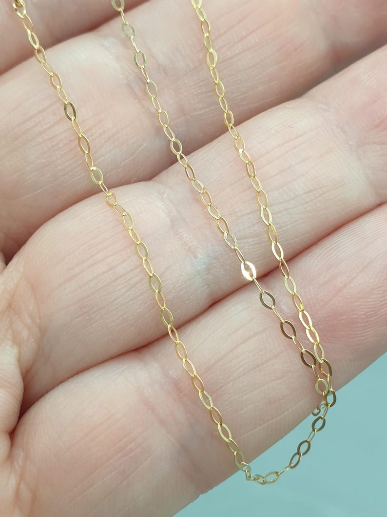 May include: A delicate gold chain necklace featuring a series of small, elongated oval links connected in a continuous pattern. The metal has a bright, reflective finish that catches the light, creating a subtle shimmer along the length of the chain.