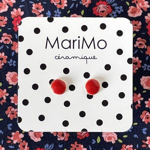 Puede incluir: Un par de pendientes de botón pequeños, redondos y de cerámica roja brillante montados sobre una tarjeta de presentación blanca con un patrón de lunares negros. La tarjeta muestra el texto MariMo céramique en letras negras sobre los pendientes.