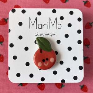 Può includere: Una ciliegia in ceramica rossa con una foglia verde e un viso sorridente. La ciliegia è su una carta bianca con pois neri e il testo "MariMo céramique".