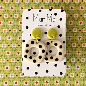May include: A pair of ceramic earrings with a cream-colored square design featuring black polka dots and a yellow circle on top. The earrings are displayed on a white card with the text "MariMo céramique" and black polka dots, set against a green floral background.