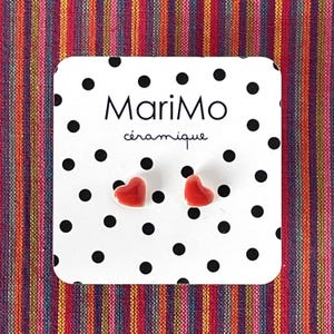 Puede incluir: Un par de pequeños pendientes de botón de cerámica roja brillante en forma de corazón, sujetos a una tarjeta expositora blanca con un patrón de lunares negros y el texto MariMo céramique impreso en letras negras en la parte superior.